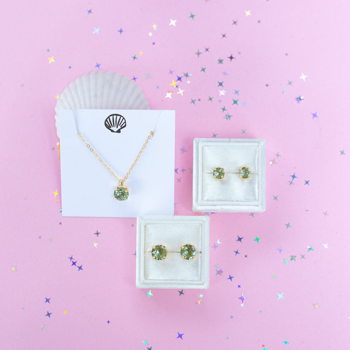 A gold chain necklace with a round Olive green pendant, displayed on a white card with a gold seashell logo, set against a pink background with star confetti.