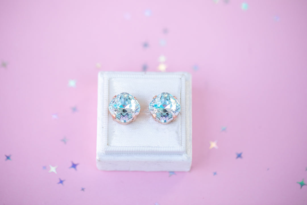 Pair of square-shaped crystal earrings on a white stand against a pink background with starbursts.