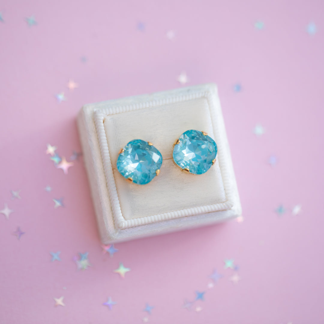 Blue gemstone earrings on a white cushion against a pink background with star-shaped decorations.