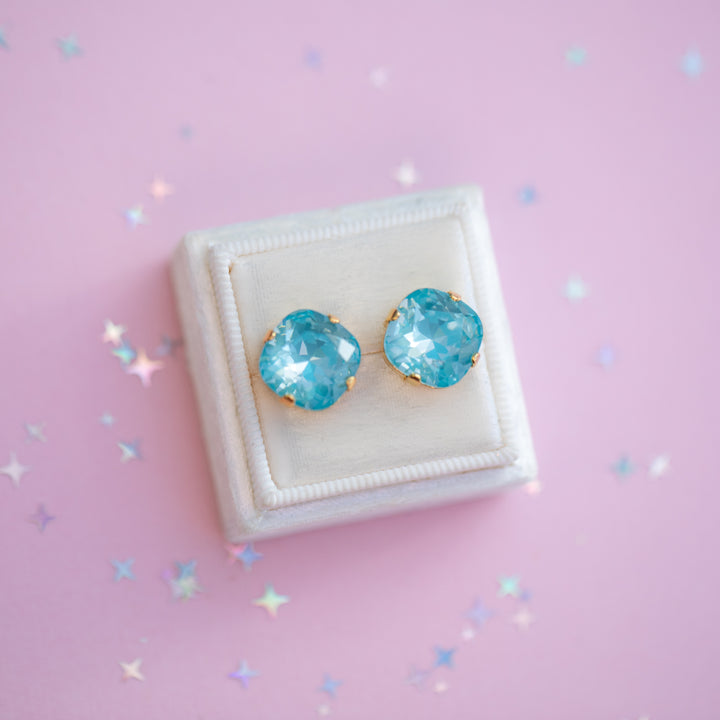 Blue gemstone earrings on a white cushion against a pink background with star-shaped decorations.