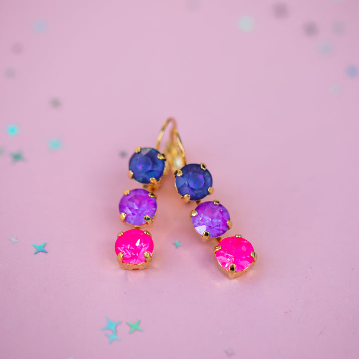 Multicolored gemstone earrings on a pink background with star-shaped confetti.