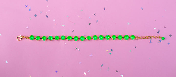 A gold chain bracelet with bright green faceted beads, laid horizontally on a pink background with scattered silver star confetti.