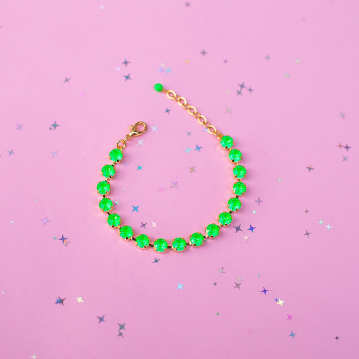 A gold chain bracelet adorned with vibrant green faceted gemstones, arranged in a curved line on a pink background scattered with small silver star confetti.