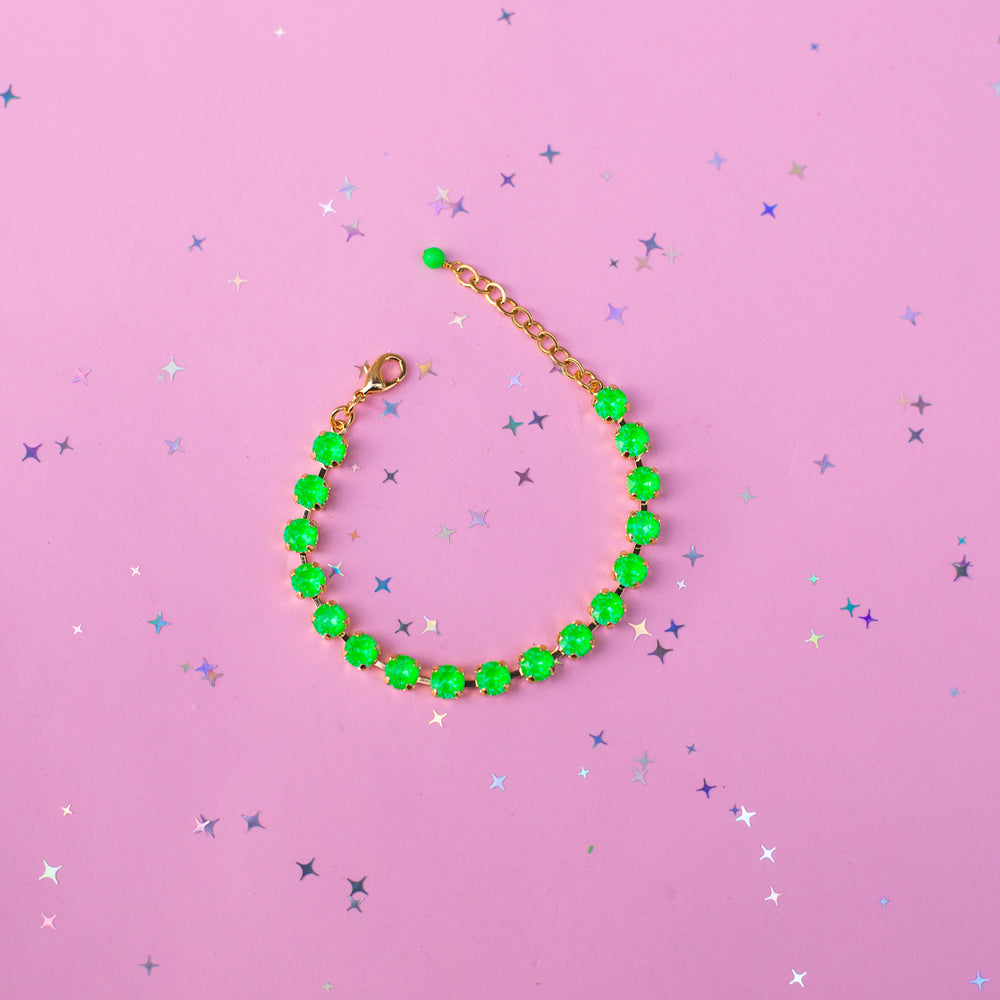 A beaded bracelet with bright green faceted beads and a gold-tone chain extender, displayed on a pink background with scattered silver star confetti.