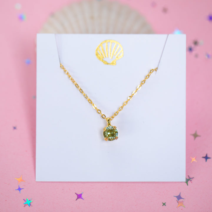 A gold chain necklace with a small olive green crystal pendant on a white card.