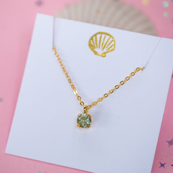 A gold chain necklace with a small olive green crystal pendant on a white card.