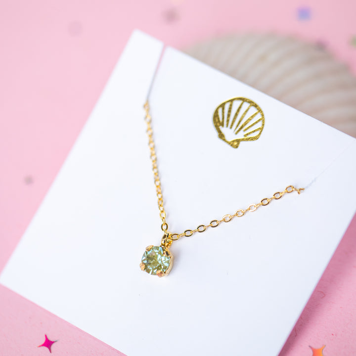A gold chain necklace with a small olive green crystal pendant on a white card.
