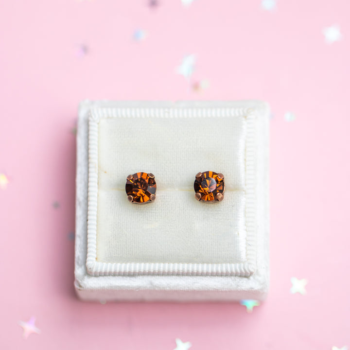 A pair of round gold stud earrings with amber-orange marquise-cut crystals, displayed on a white textured jewelry box against a pink background with scattered star confetti.