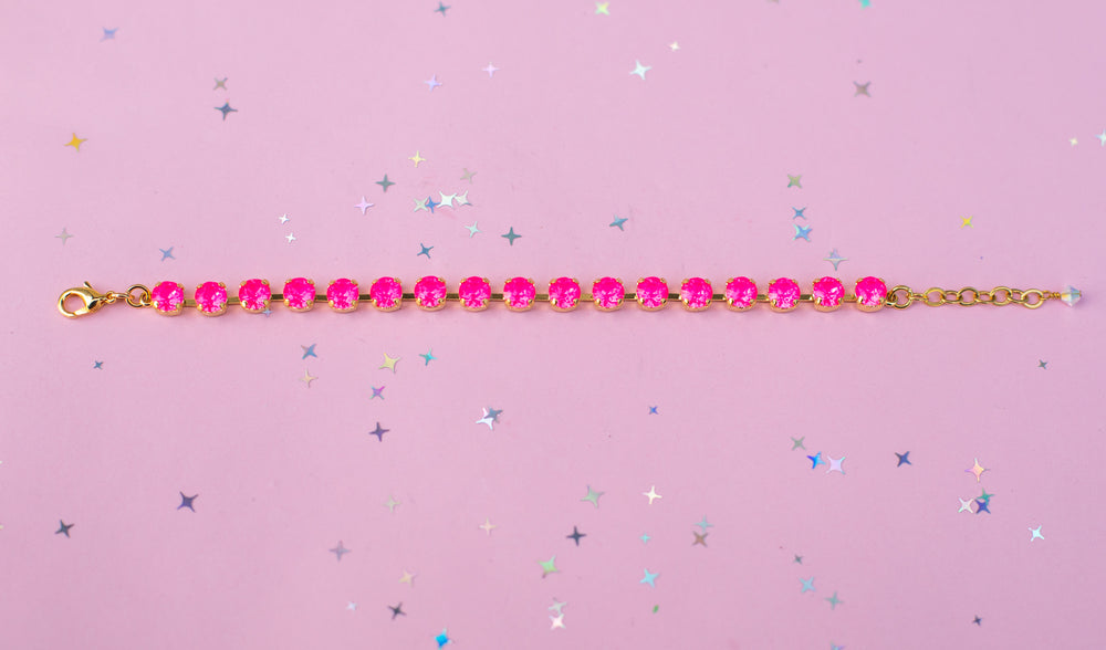 A gold chain bracelet with a row of bright pink faceted rhinestones, laid horizontally on a pink background scattered with small silver and blue star confetti.