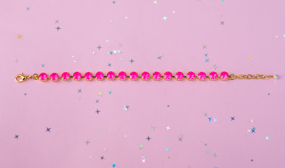 A gold chain bracelet with a row of bright pink faceted rhinestones, laid horizontally on a pink background scattered with small silver and blue star confetti.