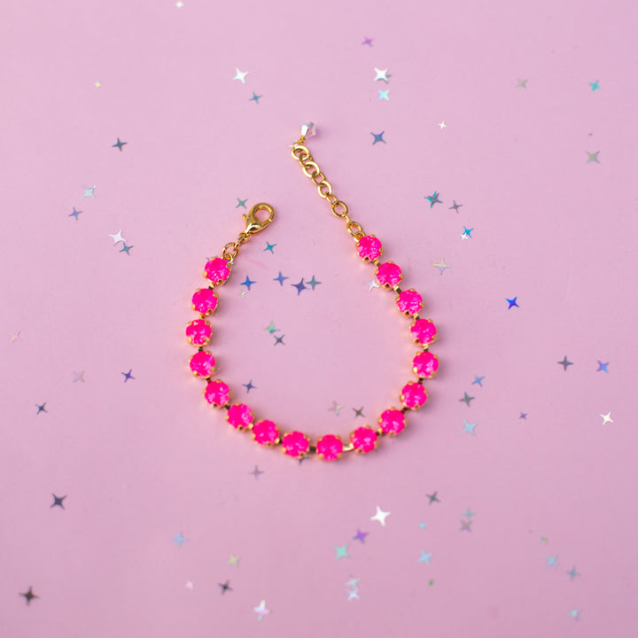 A gold chain bracelet adorned with bright hot pink faceted gemstones, arranged in a teardrop shape on a light pink background scattered with silver and blue star confetti.