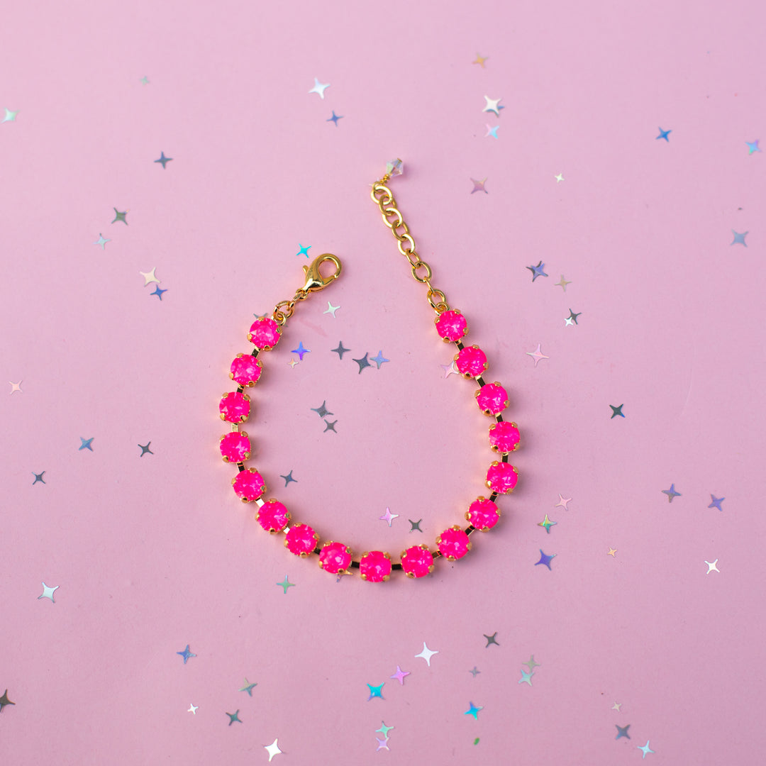 A gold-tone chain bracelet adorned with bright hot pink faceted gemstones, arranged in a curved line on a light pink background scattered with small silver and blue star confetti.