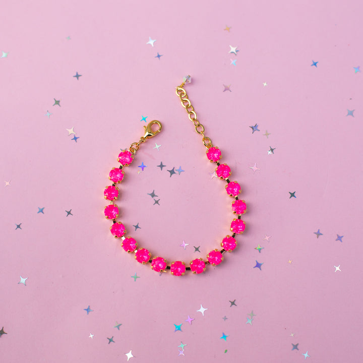 A gold-tone chain bracelet adorned with bright hot pink faceted gemstones, arranged in a curved line on a light pink background scattered with small silver and blue star confetti.