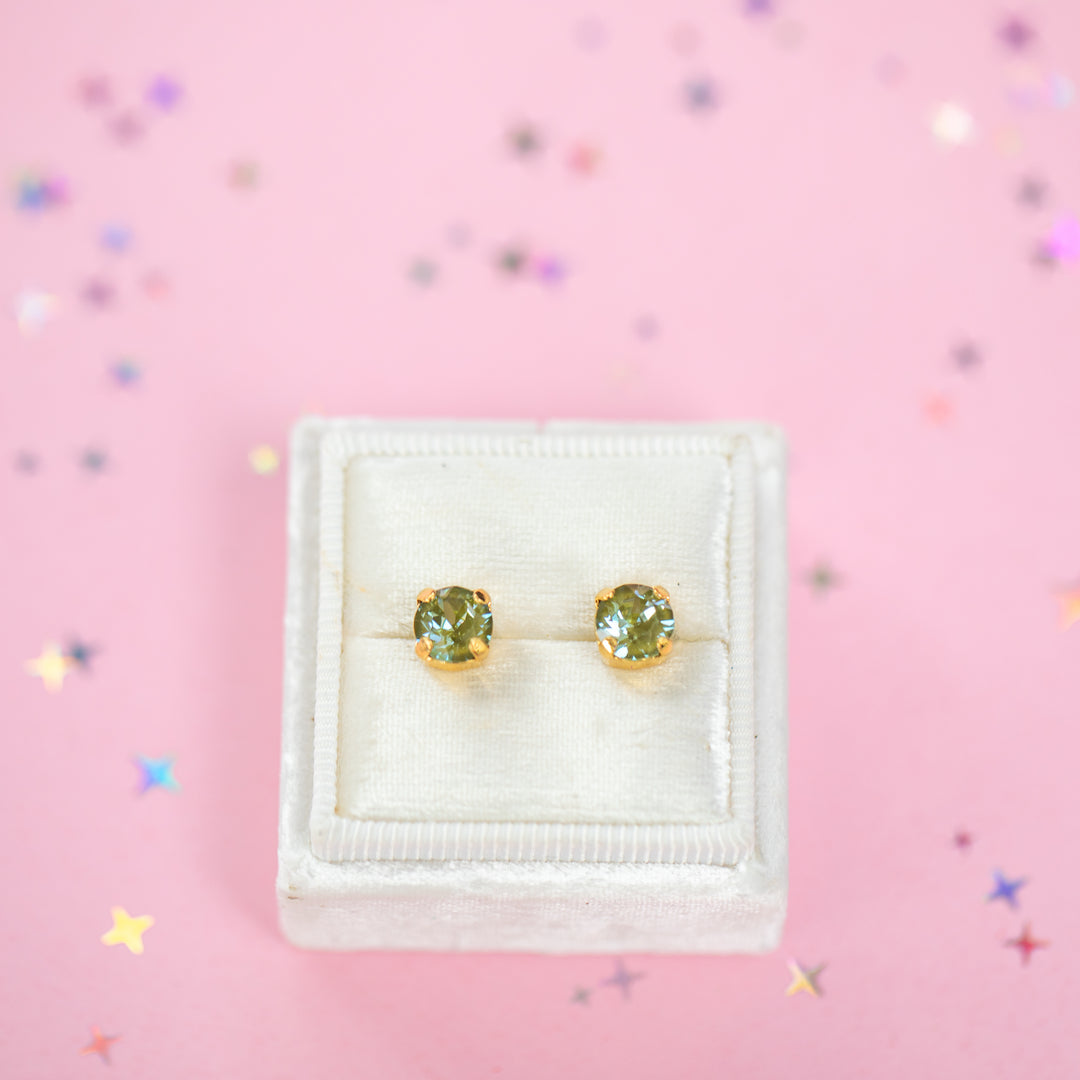A pair of gold stud earrings with round olive green gemstones, presented in a white velvet jewelry box on a pink background with scattered star confetti.