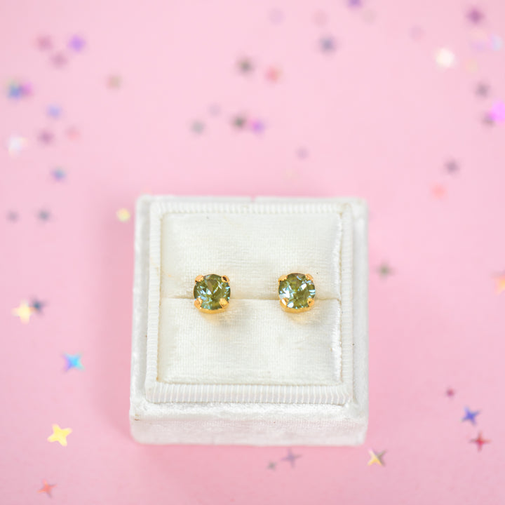 A pair of gold stud earrings with round olive green gemstones, presented in a white velvet jewelry box on a pink background with scattered star confetti.