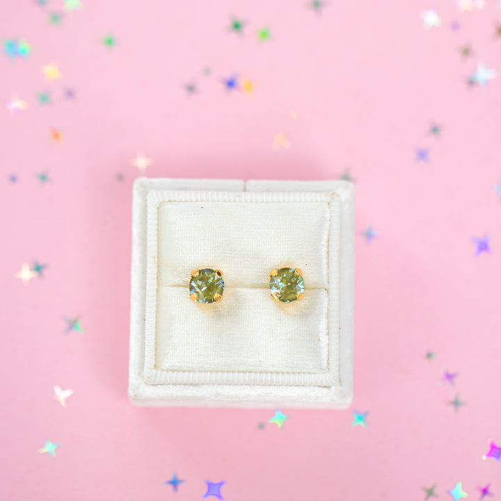 A pair of gold stud earrings with round olive green gemstones, presented in a white velvet jewelry box on a pink background with scattered star confetti.