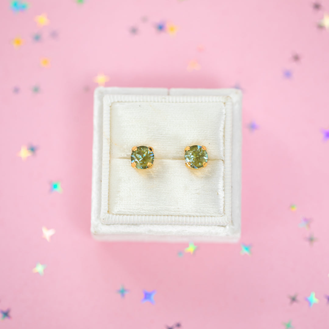 A pair of gold stud earrings with round olive green gemstones, presented in a white velvet jewelry box on a pink background with scattered star confetti.