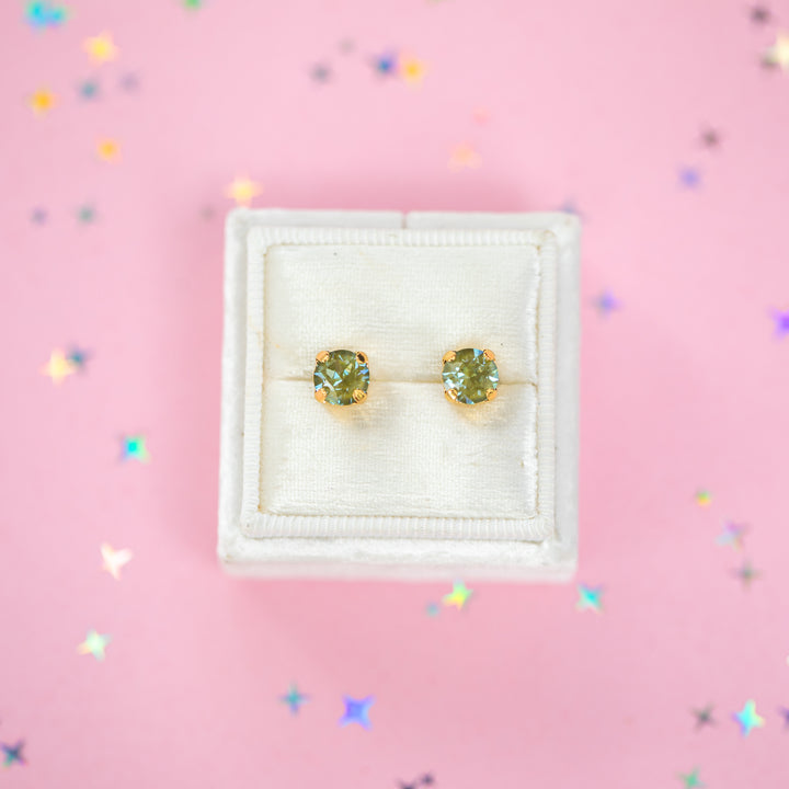 A pair of gold stud earrings with round olive green gemstones, presented in a white velvet jewelry box on a pink background with scattered star confetti.