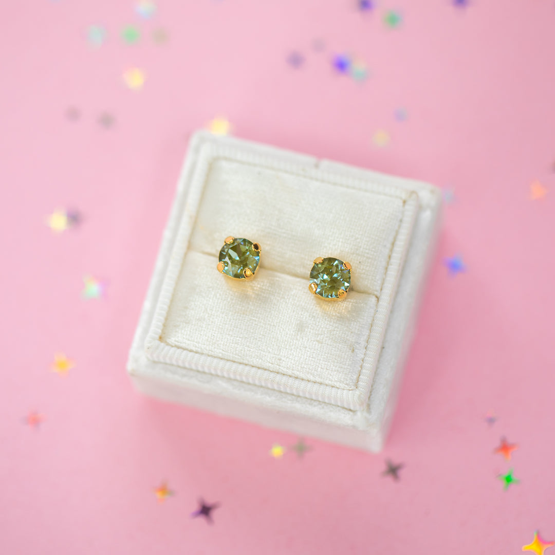 A pair of gold stud earrings with round olive green gemstones, presented in a white velvet jewelry box on a pink background with scattered star confetti.