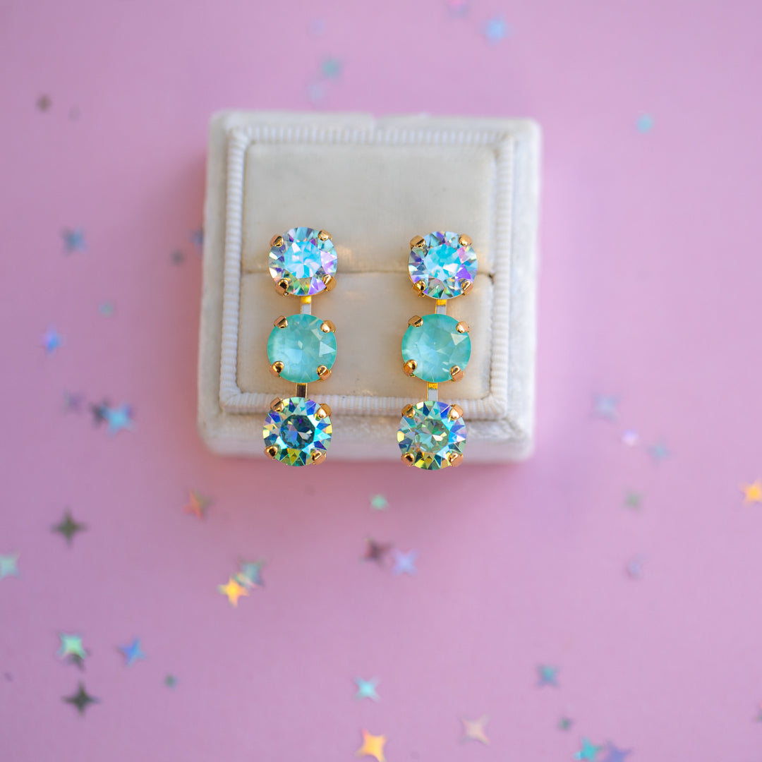 Pair of earrings with turquoise stones in a white box on a pink background with star-shaped confetti.