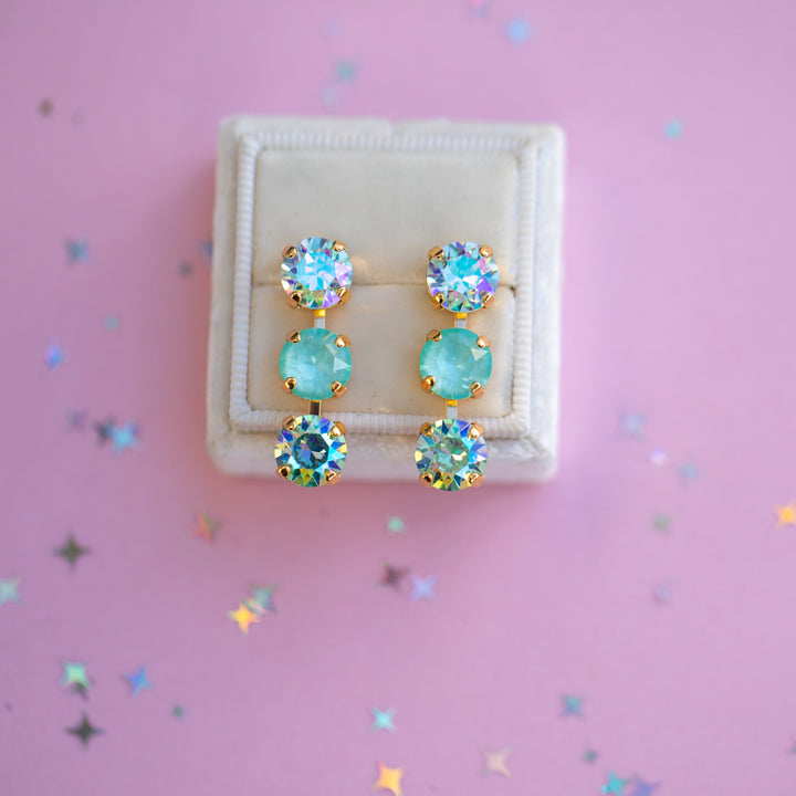 Pair of earrings with turquoise stones in a white box on a pink background with star-shaped confetti.