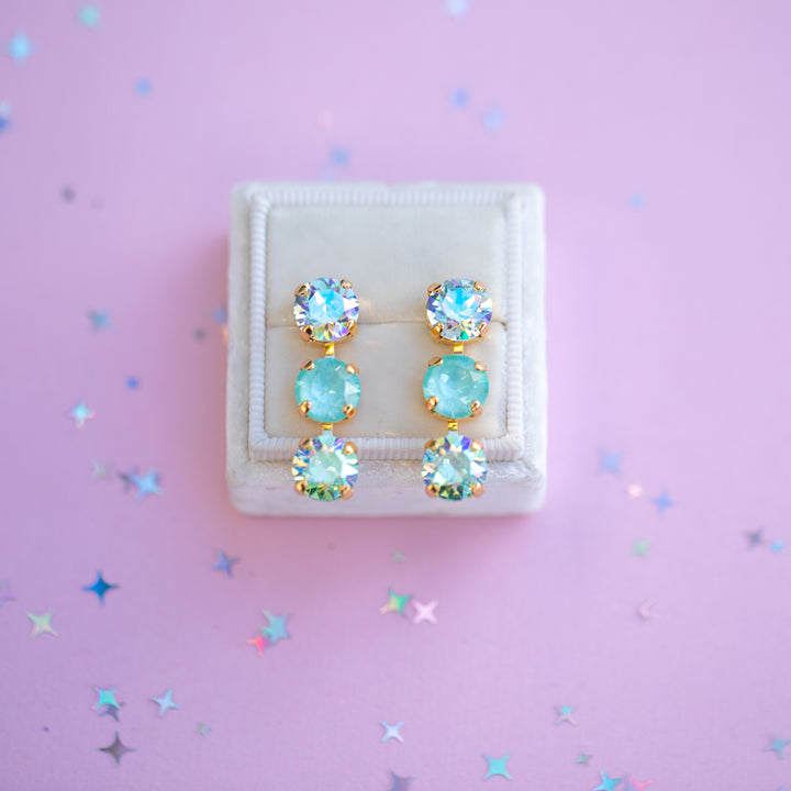 Pair of earrings with blue gemstones in a white box on a pink background with star confetti.