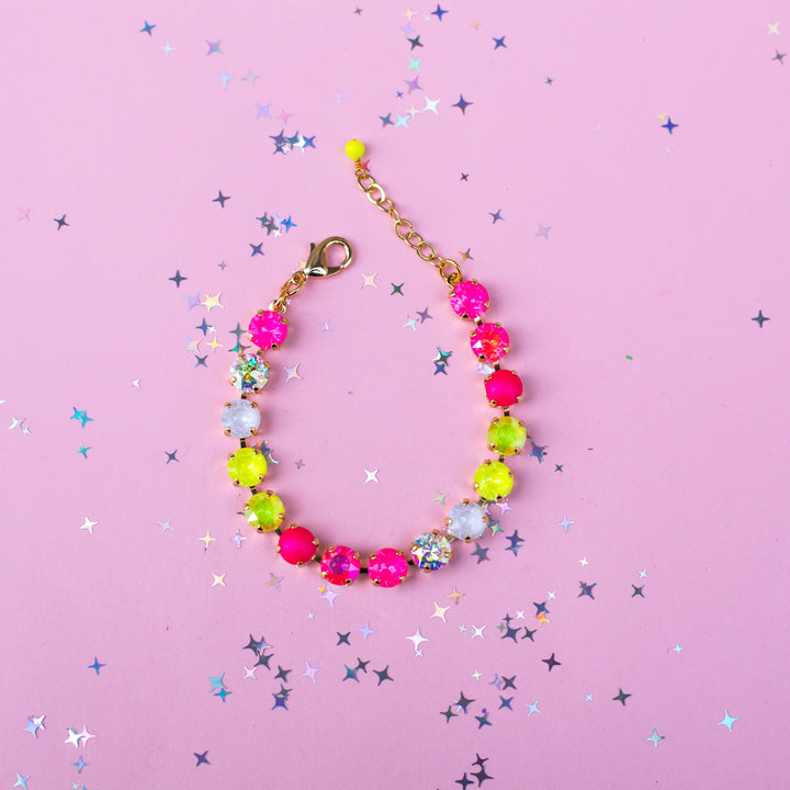 A gold chain bracelet with colorful faceted gemstone beads in hot pink, lime green, white, and glittery silver, arranged in a circle on a pink background with scattered star-shaped confetti.