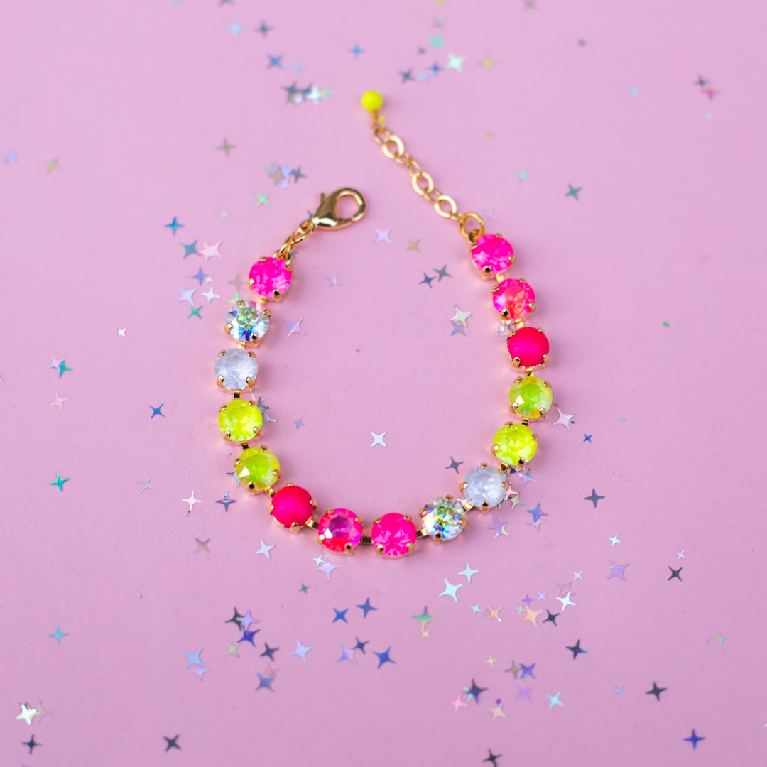 A gold-tone beaded bracelet with alternating hot pink, neon yellow, and clear faceted rhinestones, displayed on a pink background scattered with silver star confetti.