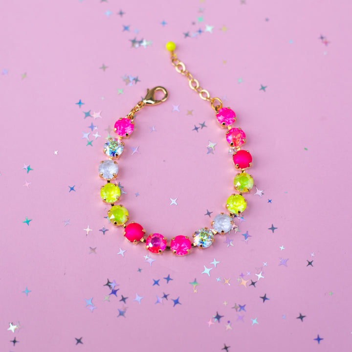 A gold-tone beaded bracelet with alternating hot pink, neon yellow, and clear faceted rhinestones, displayed on a pink background scattered with silver star confetti.
