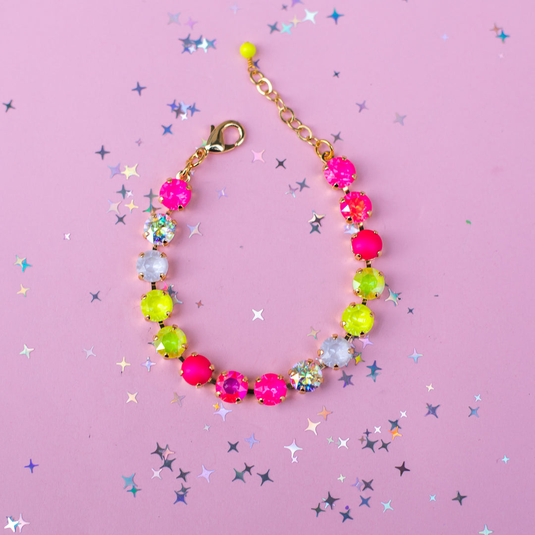 A beaded bracelet with alternating hot pink, neon yellow-green, and clear faceted rhinestones, connected by a gold-tone chain and lobster clasp, displayed on a pink background with scattered star-shaped confetti.