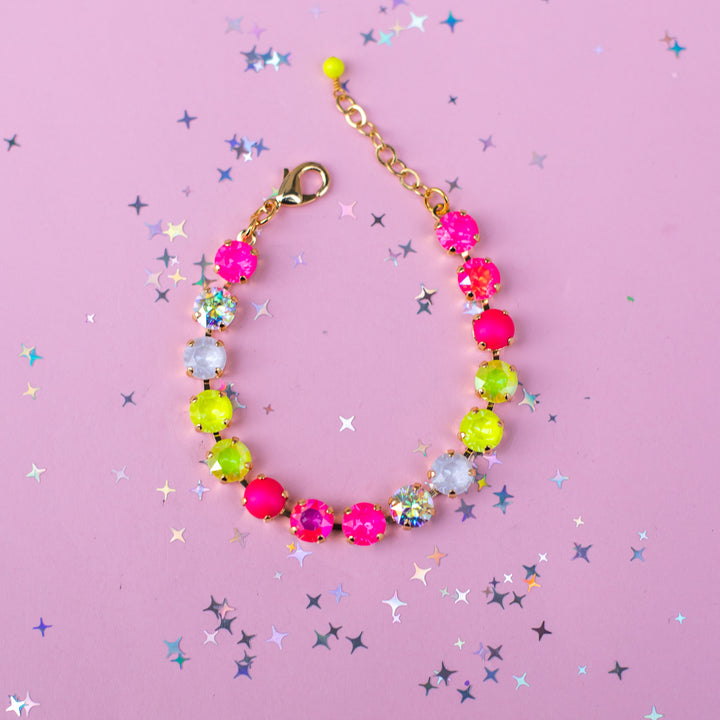 A beaded bracelet with alternating hot pink, neon yellow-green, and clear faceted rhinestones, connected by a gold-tone chain and lobster clasp, displayed on a pink background with scattered star-shaped confetti.