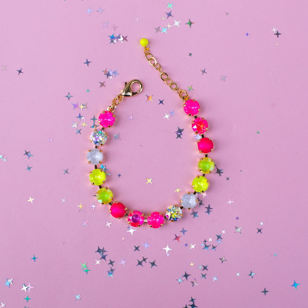 A colorful beaded bracelet with neon pink, lime green, and white faceted beads, gold-toned chain, and lobster clasp, displayed on a pink background with scattered star-shaped confetti.