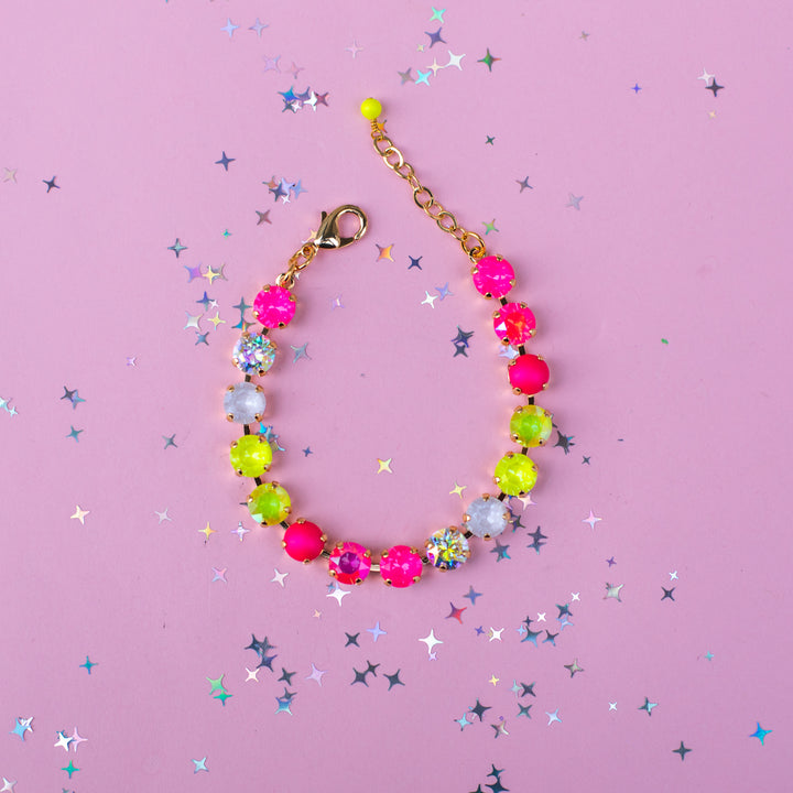 A colorful beaded bracelet with neon pink, lime green, and white faceted beads, gold-toned chain, and lobster clasp, displayed on a pink background with scattered star-shaped confetti.