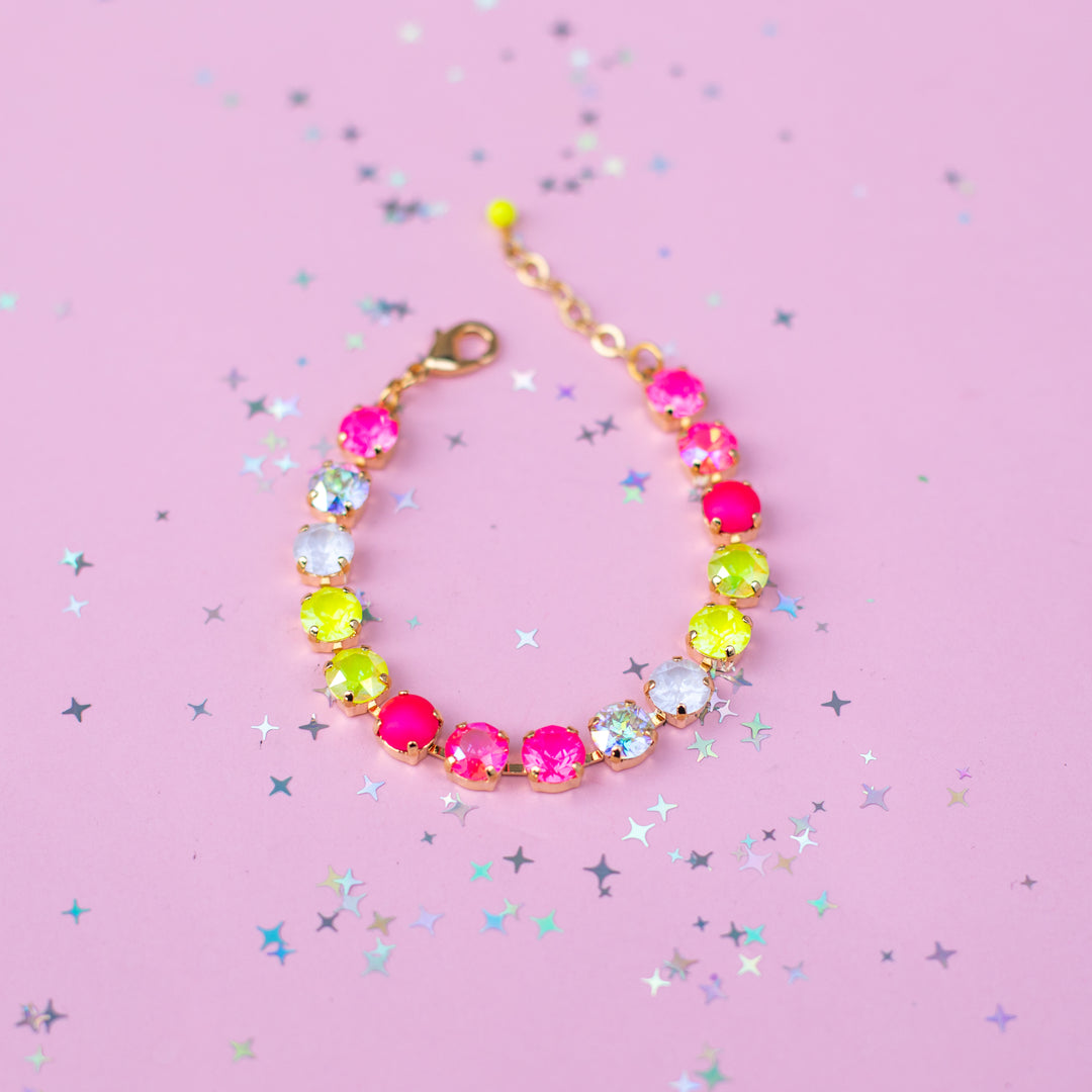 A gold-tone beaded bracelet featuring vibrant neon pink, lime green, and light blue faux gemstones, displayed on a pink background scattered with silver star confetti.