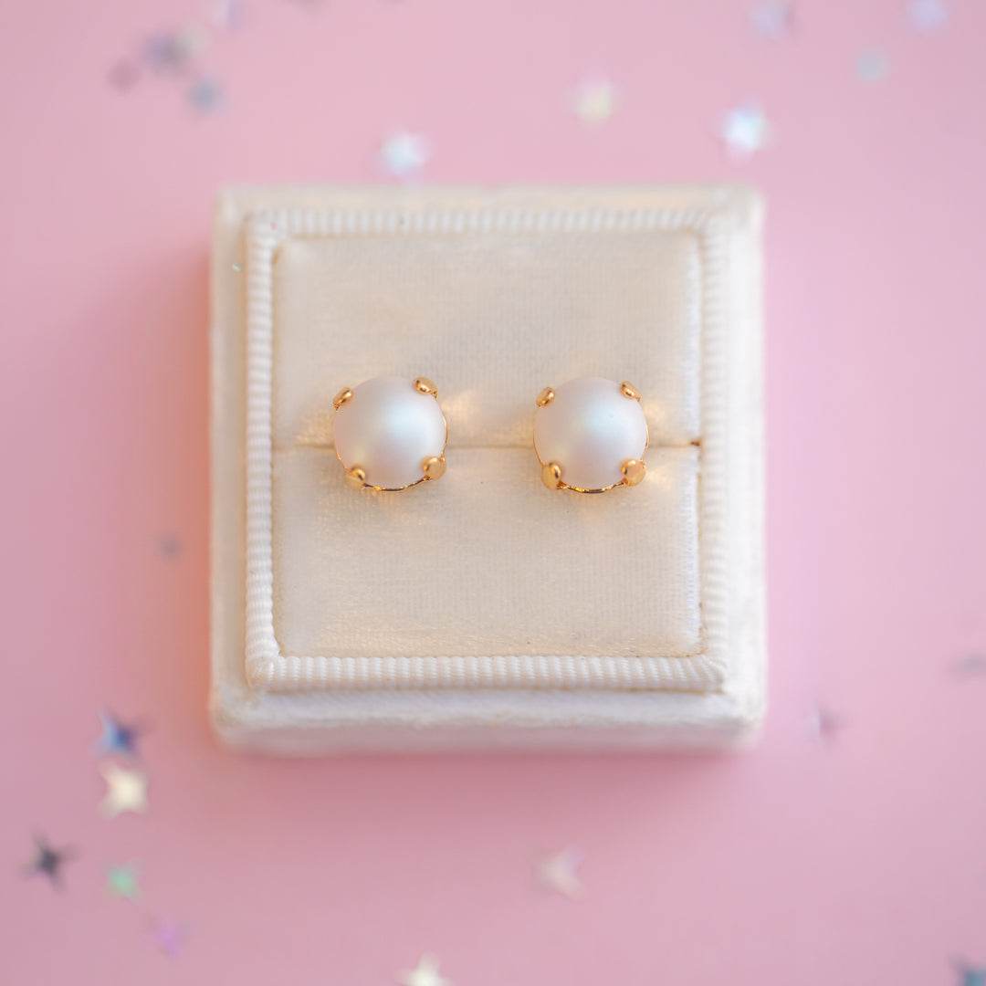 Pearl earrings with gold accents on a white cushion in a jewelry box with a pink background