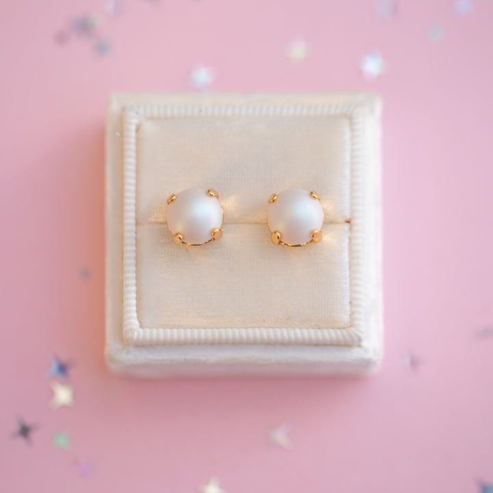 Pearl earrings with gold accents on a white cushion in a jewelry box with a pink background