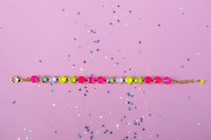 A beaded bracelet with alternating hot pink, neon yellow-green, and white faceted beads on a gold-tone chain, displayed on a pink background scattered with silver star confetti.