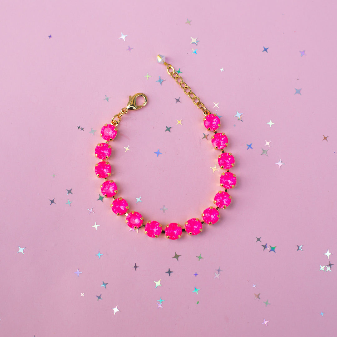 A gold chain bracelet with bright pink faceted gemstones, displayed on a pink background scattered with small silver star confetti.