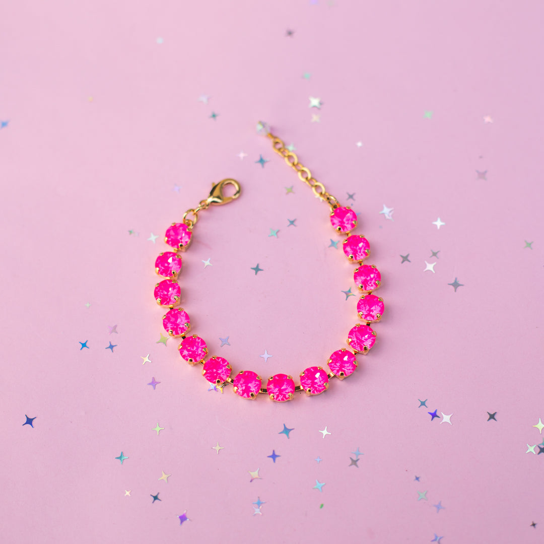 A gold chain bracelet with round, bright pink gemstones, displayed on a pink background with scattered silver star confetti.