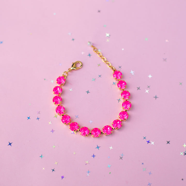 A gold chain bracelet with round, bright pink gemstones, displayed on a pink background with scattered silver star confetti.