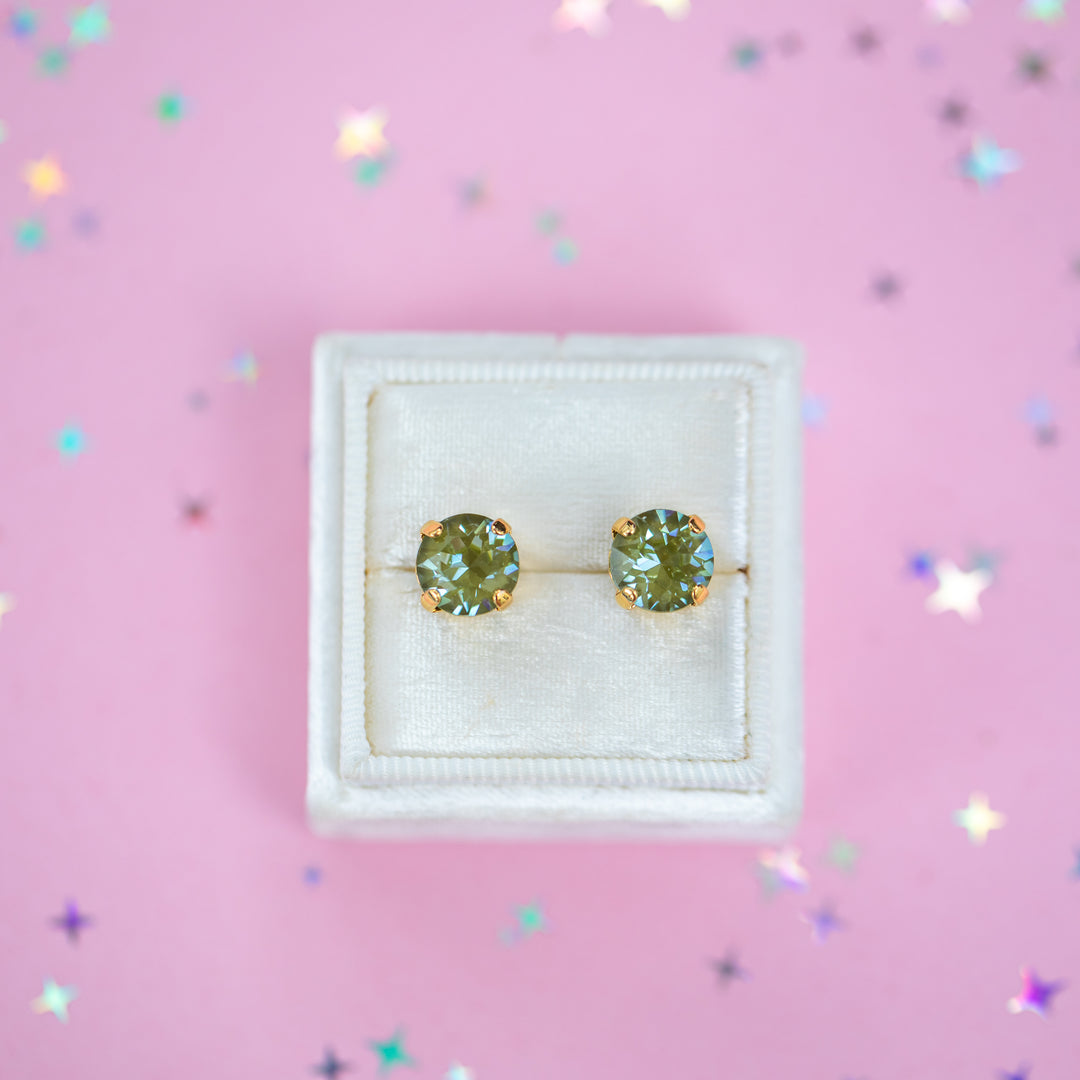 A pair of round Olive green crystal studs in a gold setting on a velvet display box with a pink background.