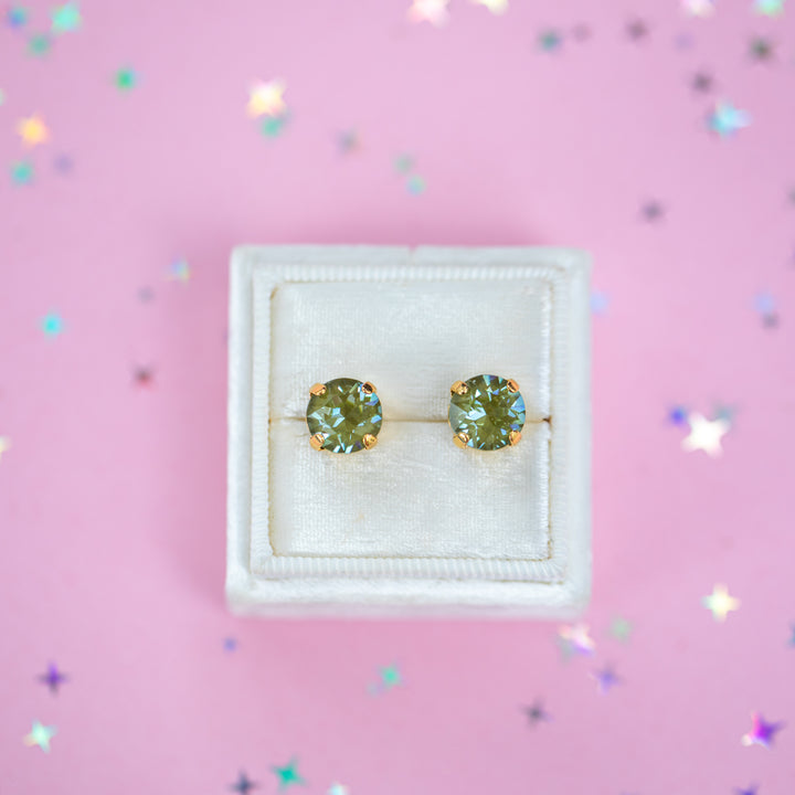 A pair of round Olive green crystal studs in a gold setting on a velvet display box with a pink background.