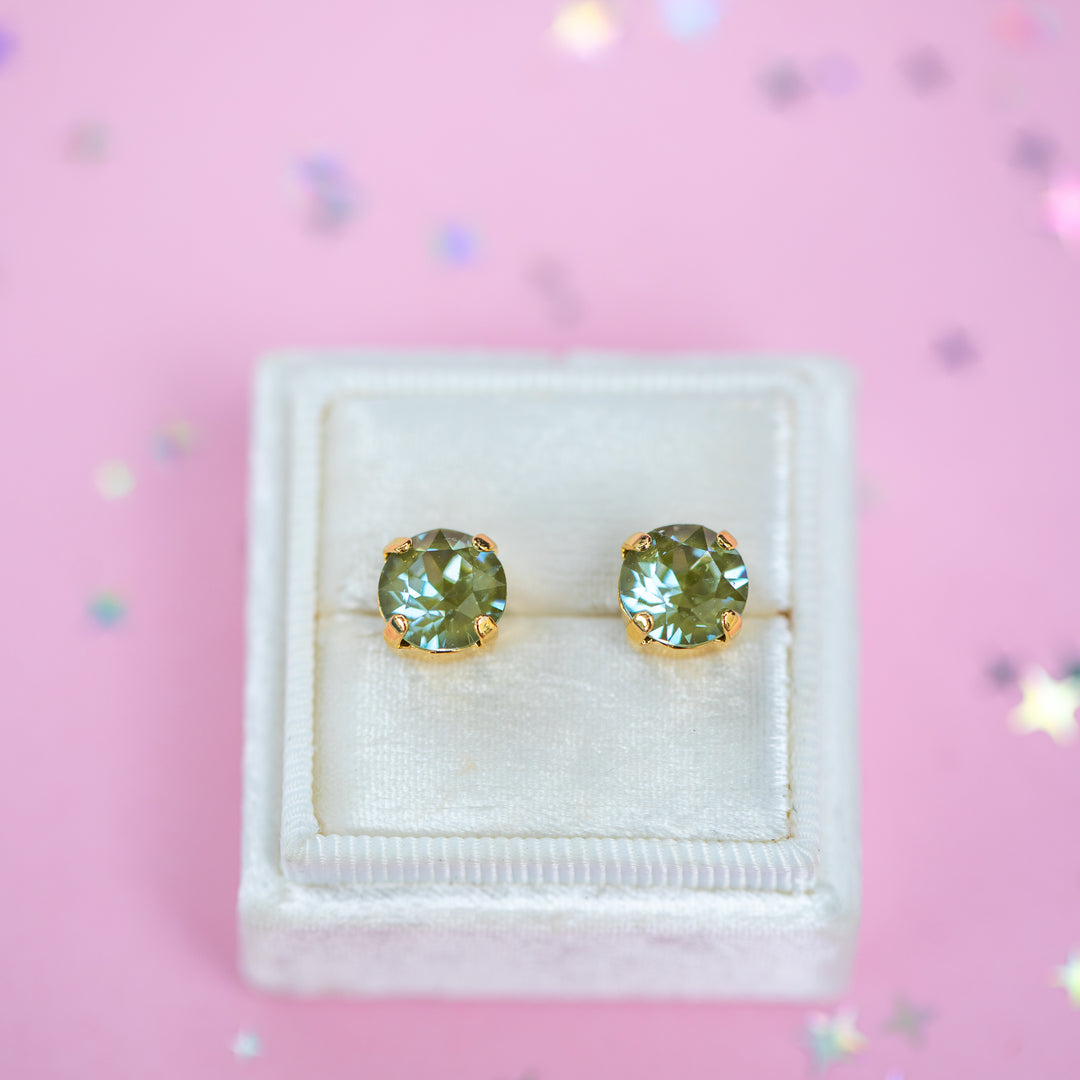 A pair of round Olive green crystal studs in a gold setting on a velvet display box with a pink background.