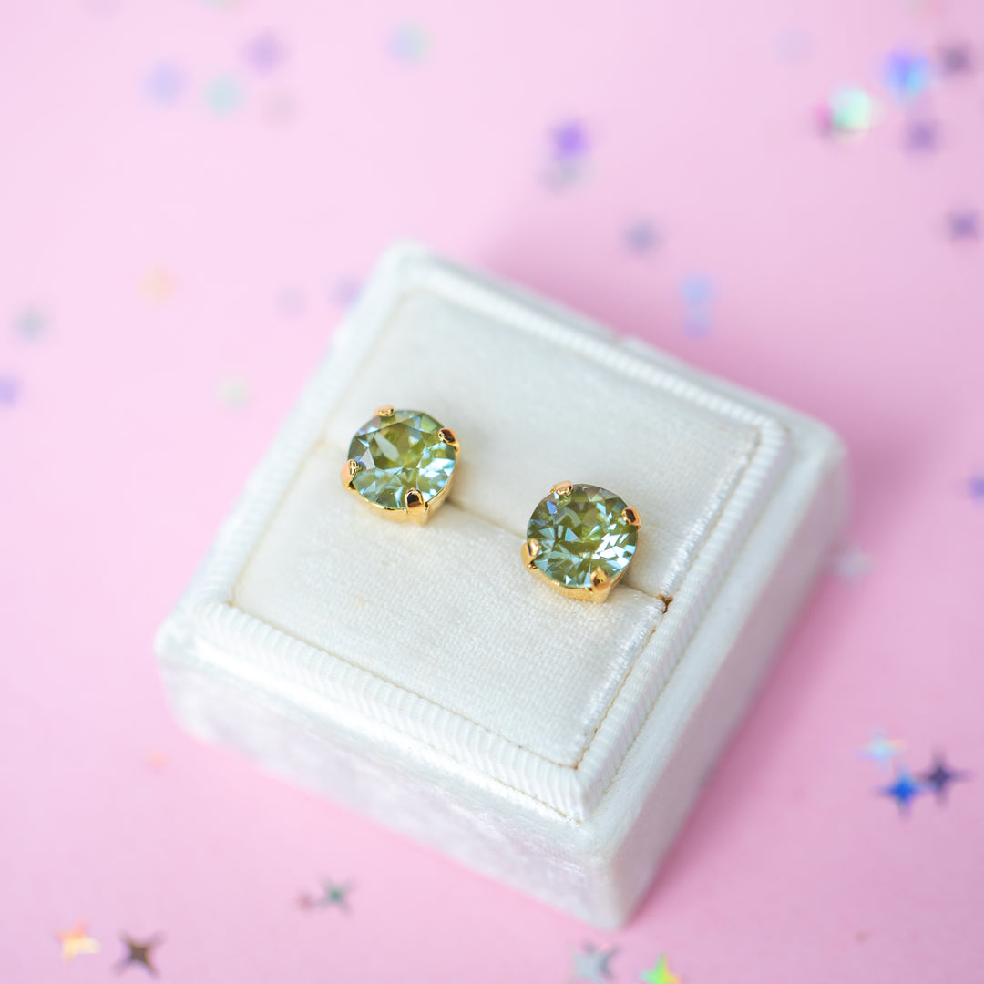 A pair of round Olive green crystal studs in a gold setting on a velvet display box with a pink background.