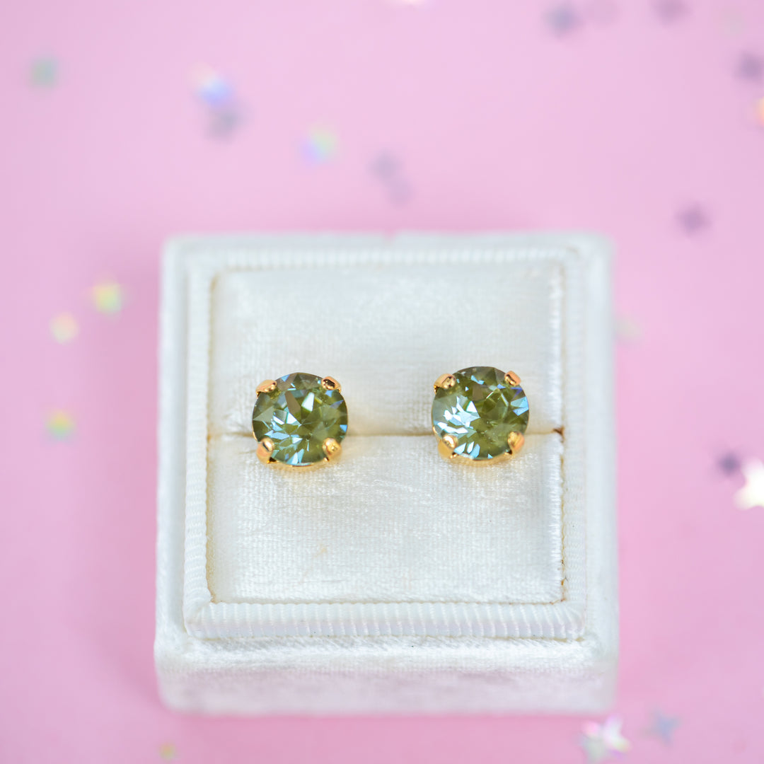 A pair of round Olive green crystal studs in a gold setting on a velvet display box with a pink background.