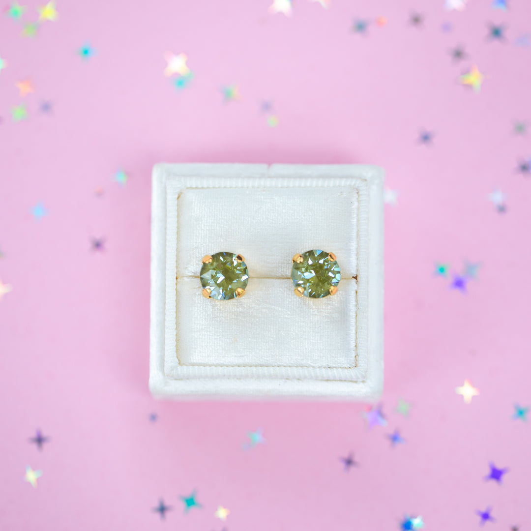 A pair of round Olive green crystal studs in a gold setting on a velvet display box with a pink background.