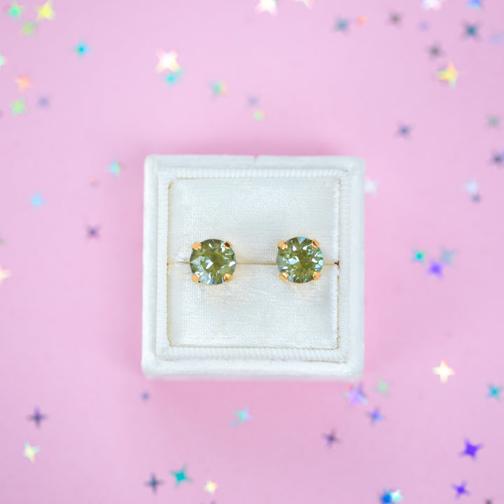 A pair of round Olive green crystal studs in a gold setting on a velvet display box with a pink background.