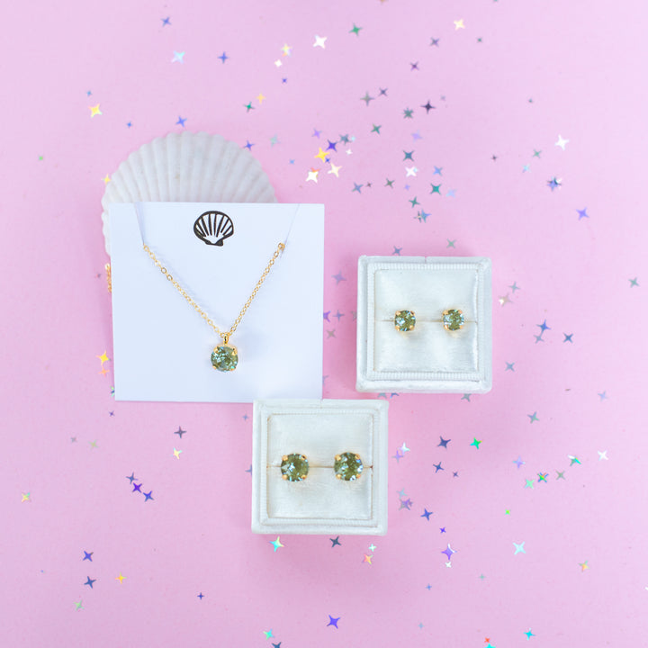 A gold chain necklace with a round Olive green pendant, displayed on a white card with a gold seashell logo, set against a pink background with star confetti.