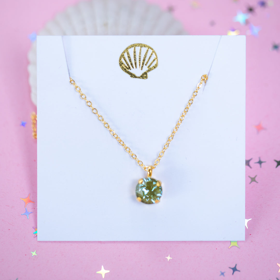 A gold chain necklace with a round Olive green pendant, displayed on a white card with a gold seashell logo, set against a pink background with star confetti.