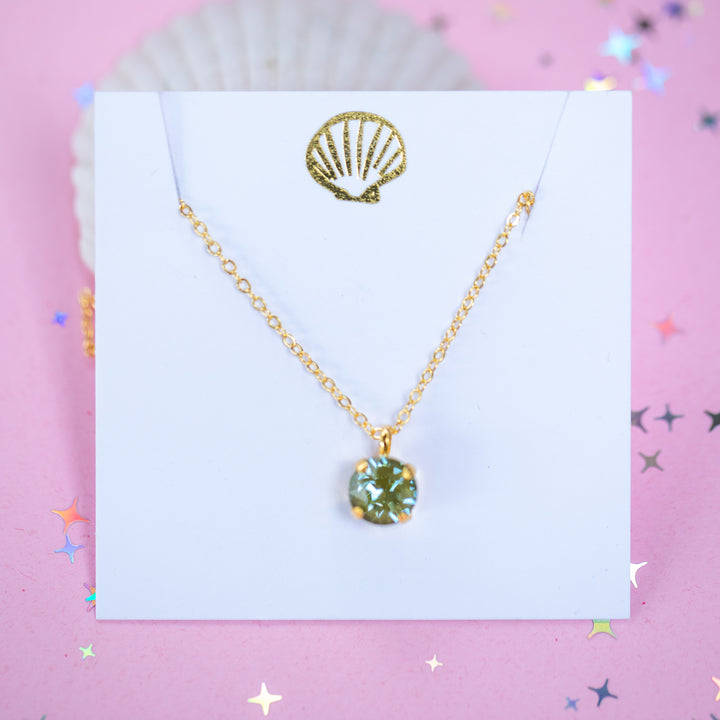 A gold chain necklace with a round Olive green pendant, displayed on a white card with a gold seashell logo, set against a pink background with star confetti.